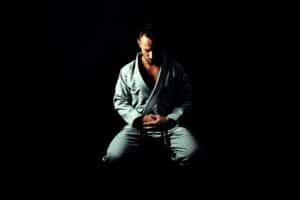 A man in a karate outfit sits on a chair in a dark environment. His head is lowered. He is concentrating